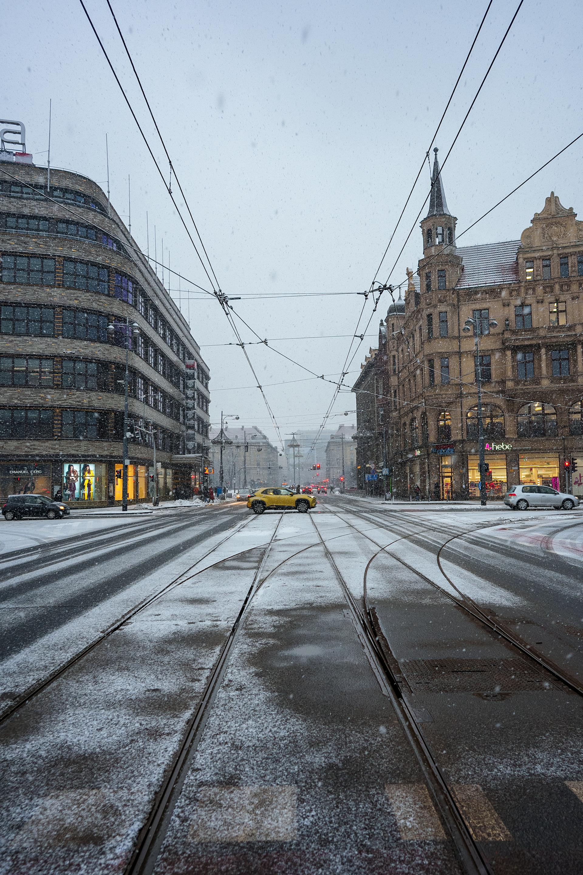 Ośnieżona miejska ulica z torami tramwajowymi prowadzącymi w dal pod zachmurzonym niebem.