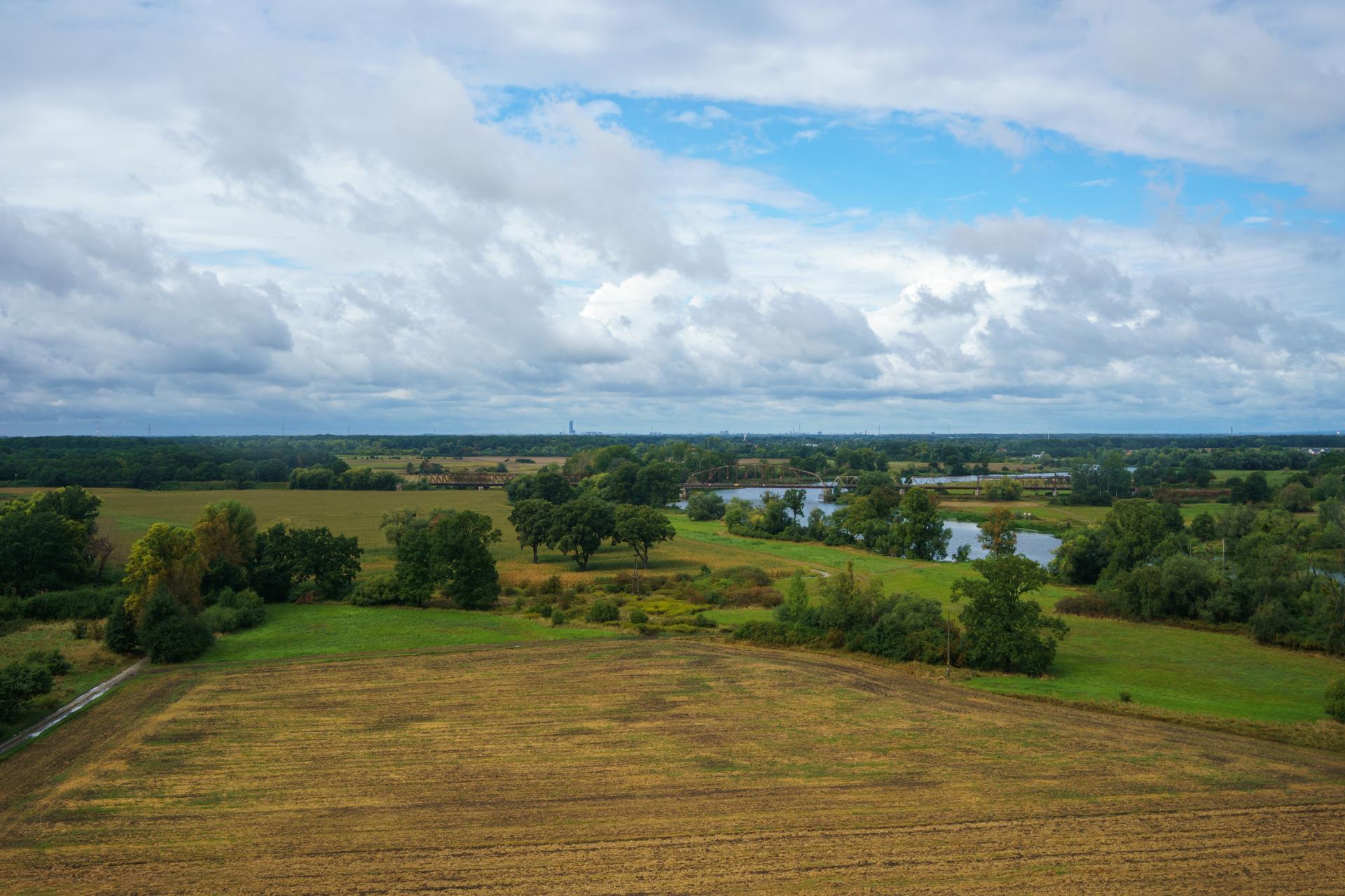 Malowniczy widok na wiejski krajobraz z rozległymi polami, kępami drzew i zakolem rzeki pod dynamicznym, zachmurzonym niebem.