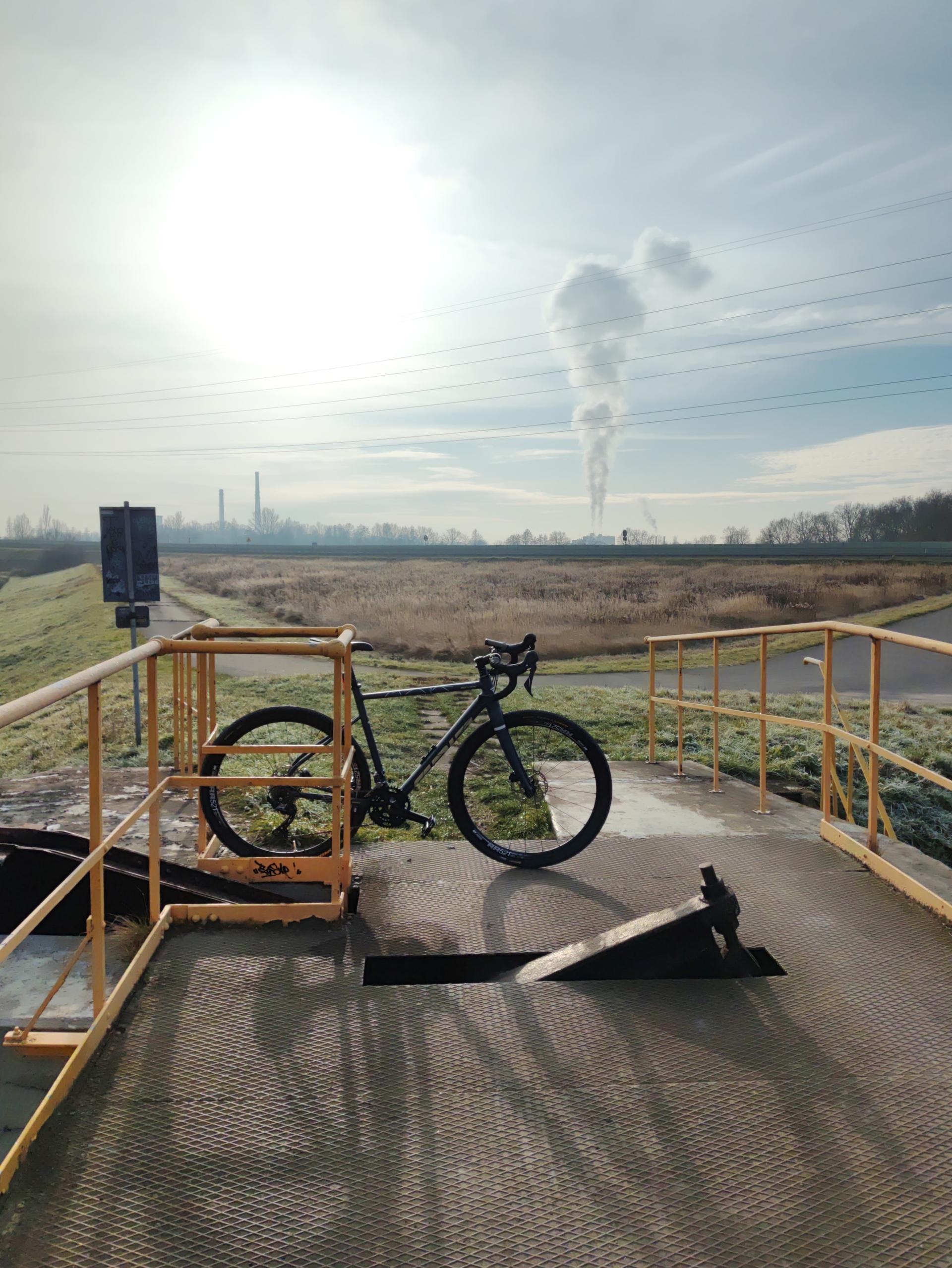 Rower gravelowy zaparkowany na betonowym podeście z widokiem na industrialny krajobraz i dymiące kominy w mroźny, słoneczny dzień.