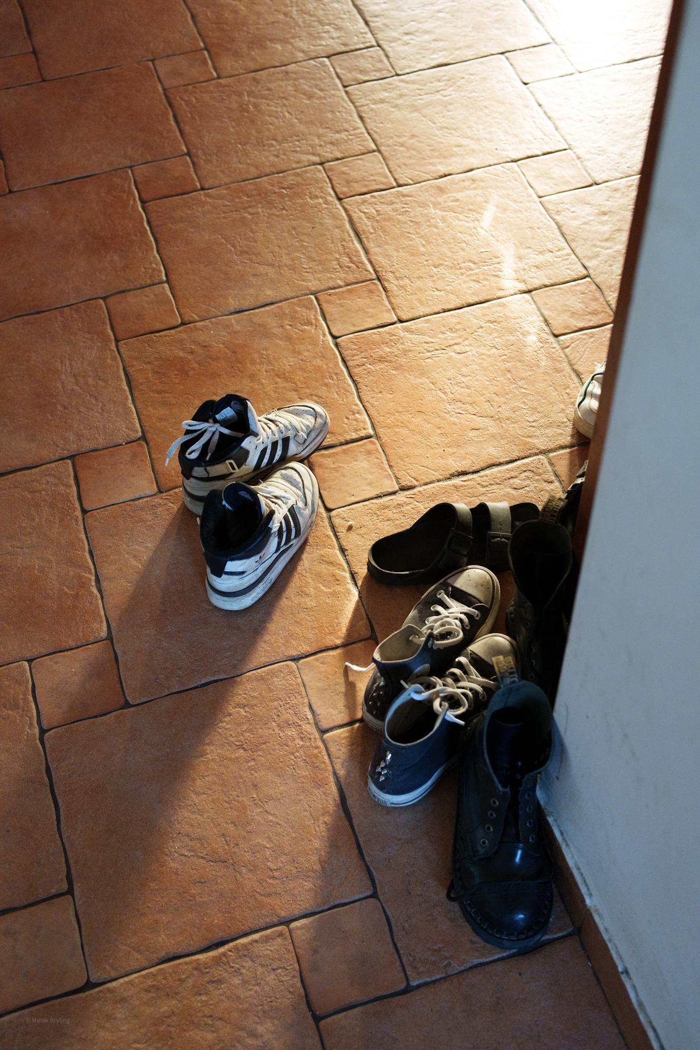 Para biało-czarnych sneakersów i kilka innych ciemnych butów pozostawionych na kafelkowej podłodze przy wejściu, oświetlonych jasnym porannym słońcem.