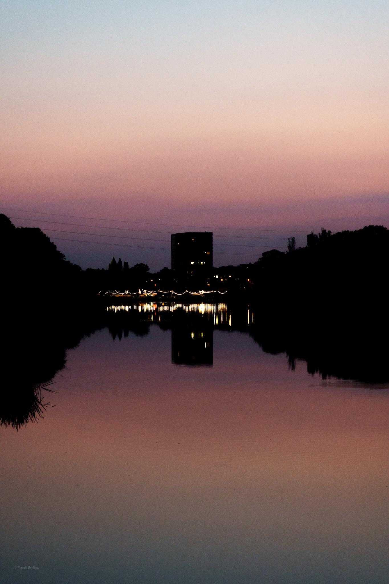 Malowniczy zachód słońca nad spokojną wodą, z sylwetką budynku odbijającą się w tafli jeziora. Niebo mieni się odcieniami różu i fioletu, tworząc spokojną i nostalgiczną atmosferę.