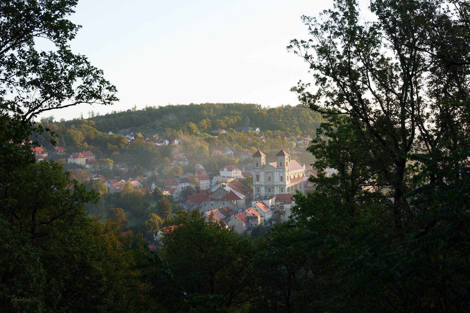 Malowniczy widok na miasteczko położone wśród zalesionych wzgórz, z charakterystyczną białą architekturą, uchwycony o poranku zza drzew.