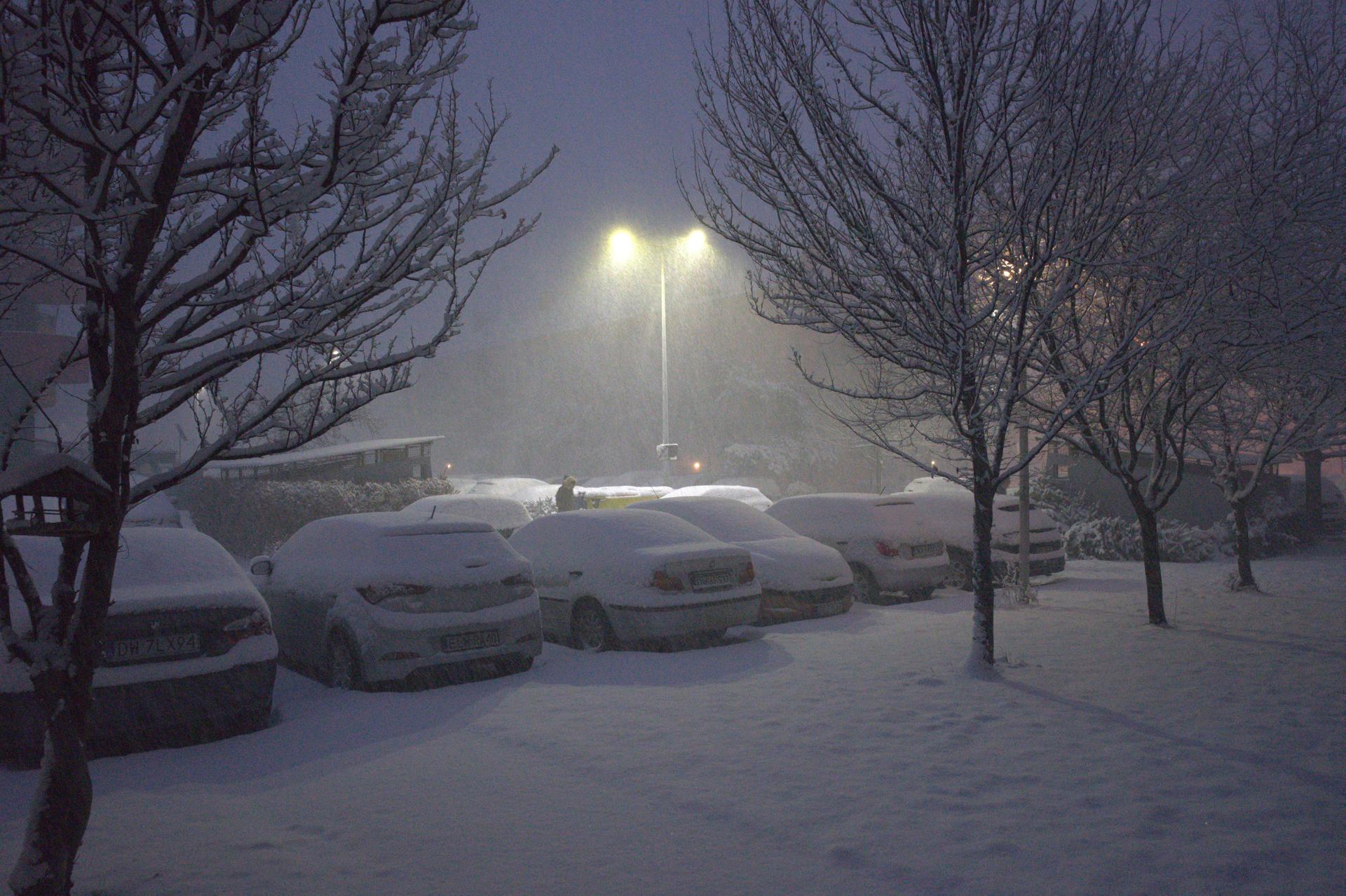 Nocny, zimowy krajobraz przedstawiający parking zasypany śniegiem. Światło latarni przebija się przez gęsto padający śnieg, oświetlając rzędy zaparkowanych aut i bezlistne drzewa.
