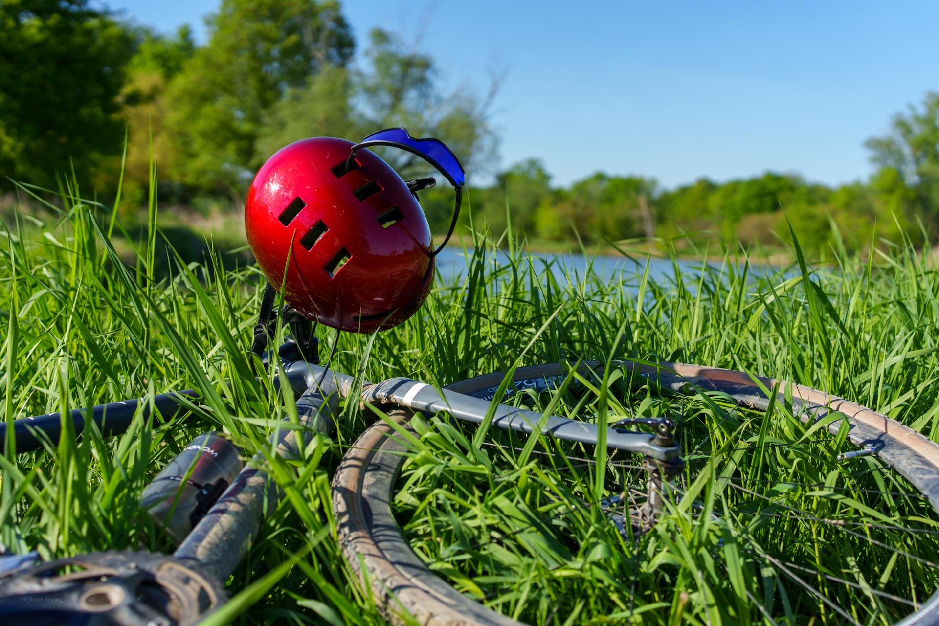 Czerwony kask rowerowy spoczywający na rowerze w gęstej, zielonej trawie nad brzegiem jeziora w słoneczny dzień.