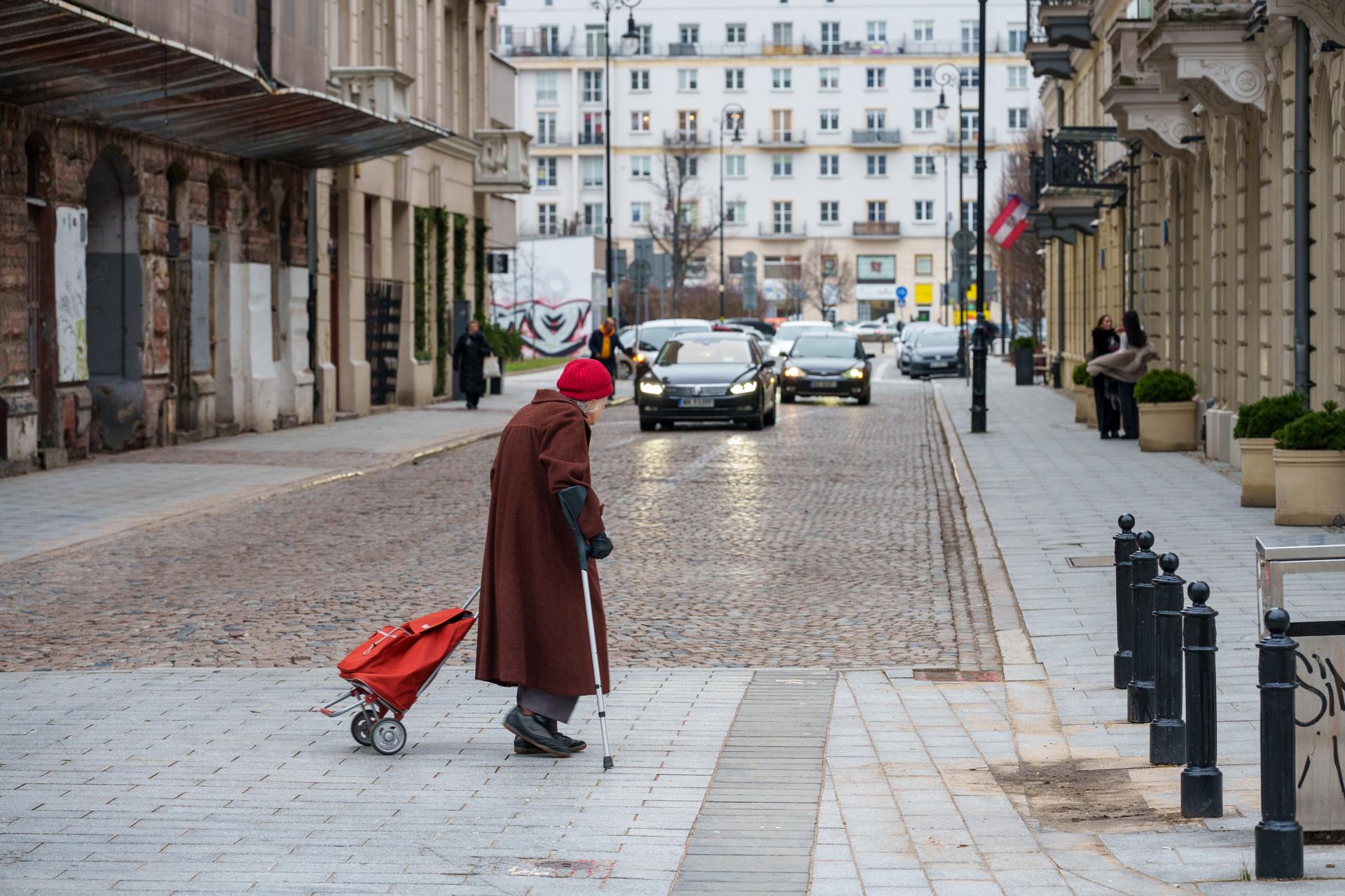 Starsza osoba w brązowym płaszczu i czerwonej czapce ciągnie czerwony wózek na zakupy po brukowanym chodniku w mieście. W tle widać ulicę z nadjeżdżającymi samochodami i kamienice.