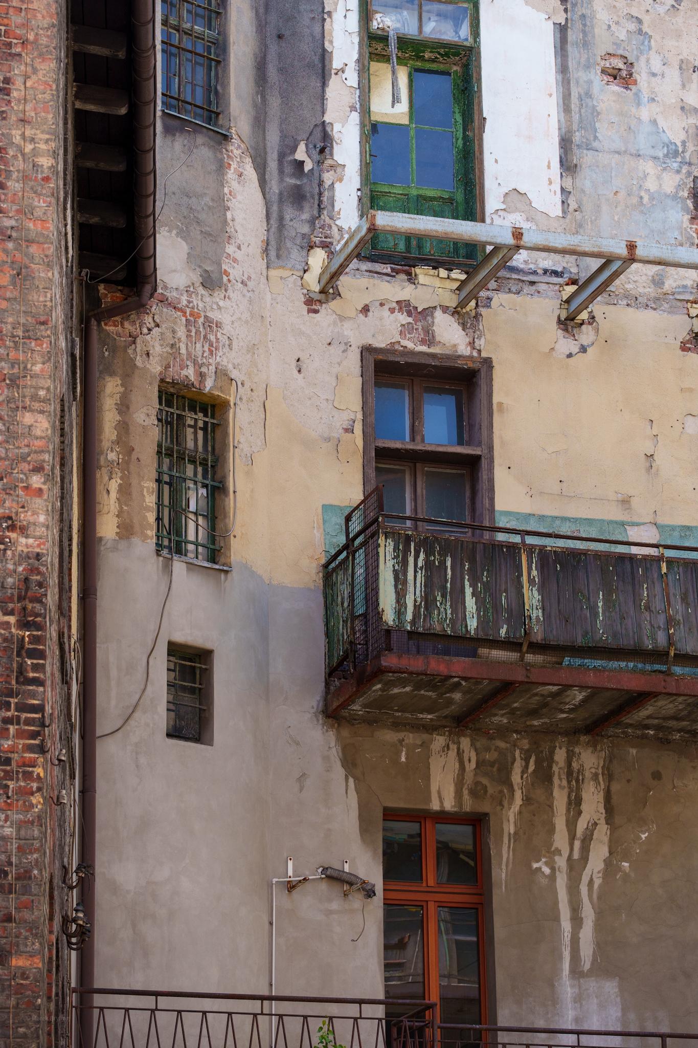 Zbliżenie na elewację starej, niszczejącej kamienicy z drewnianym balkonem i łuszczącą się farbą, ukazujące upływ czasu.
