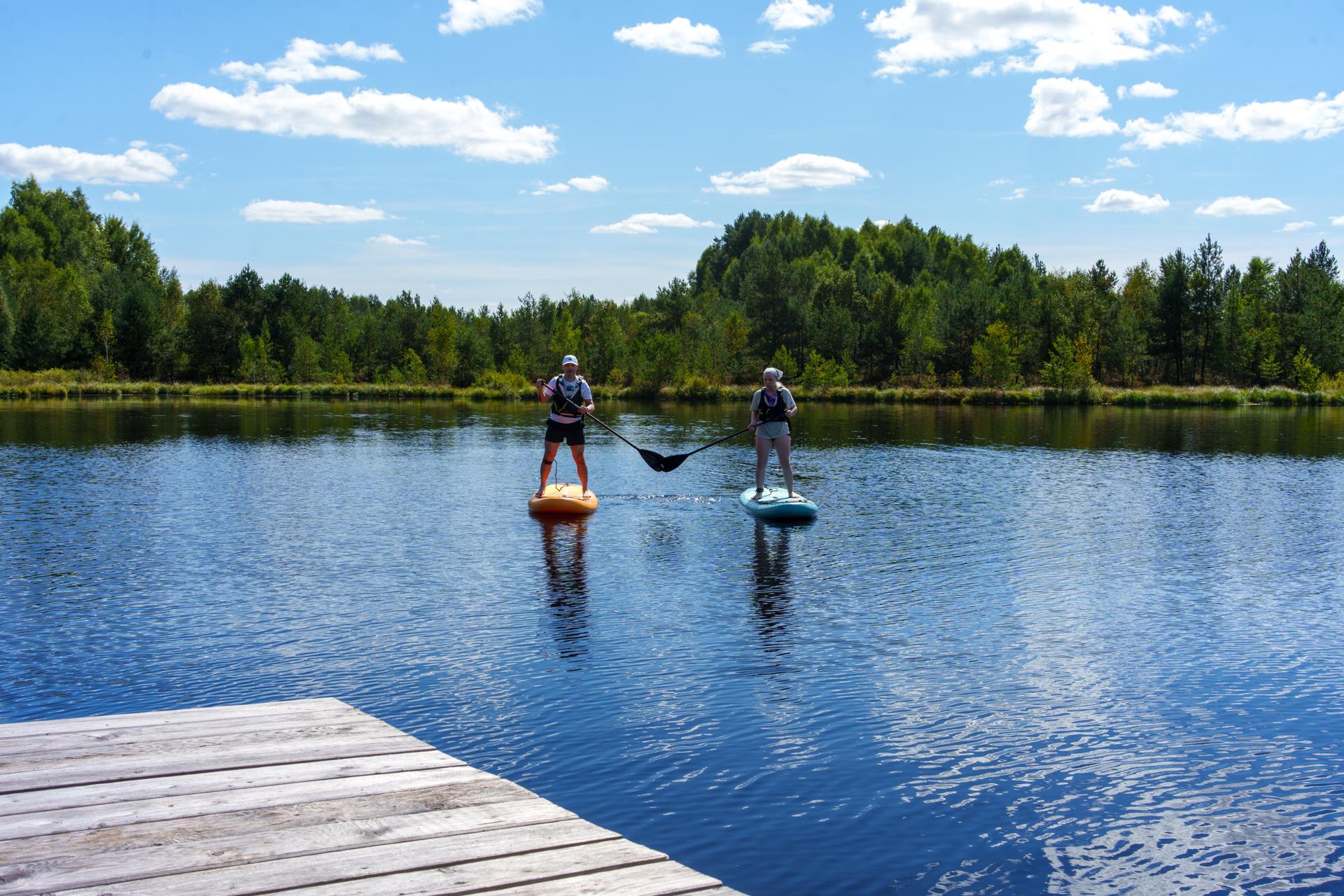 Dwoje ludzi pływających na deskach SUP na spokojnym jeziorze, otoczonych gęstym lasem pod błękitnym niebem.