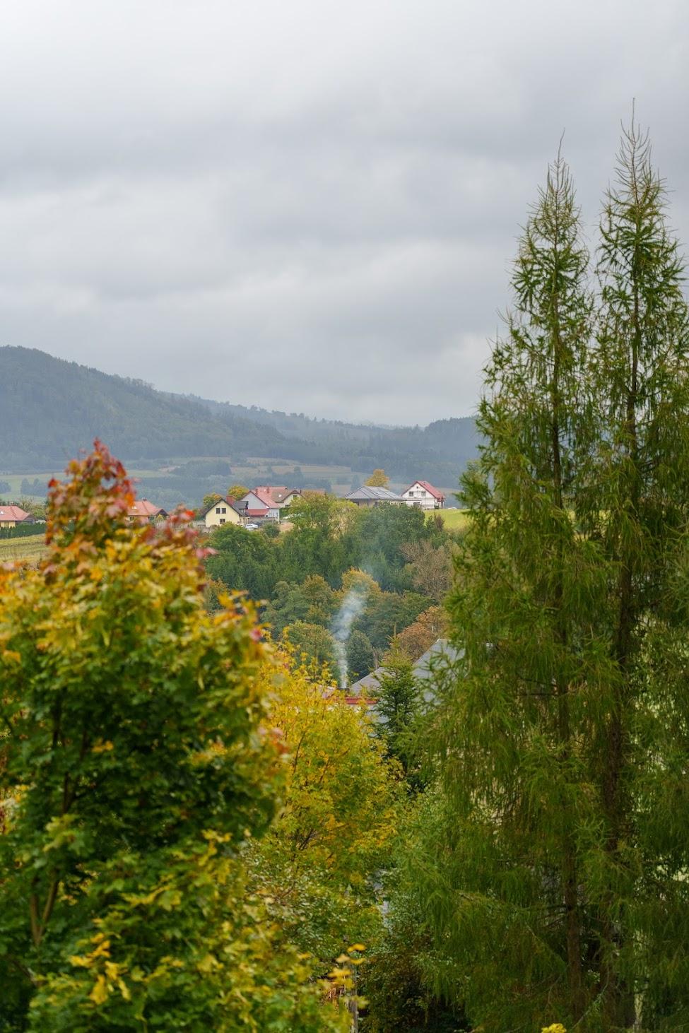 Malowniczy jesienny krajobraz z widokiem na małą miejscowość położoną u stóp wzgórz. Na pierwszym planie kolorowe drzewa w jesiennych barwach pod pochmurnym niebem.