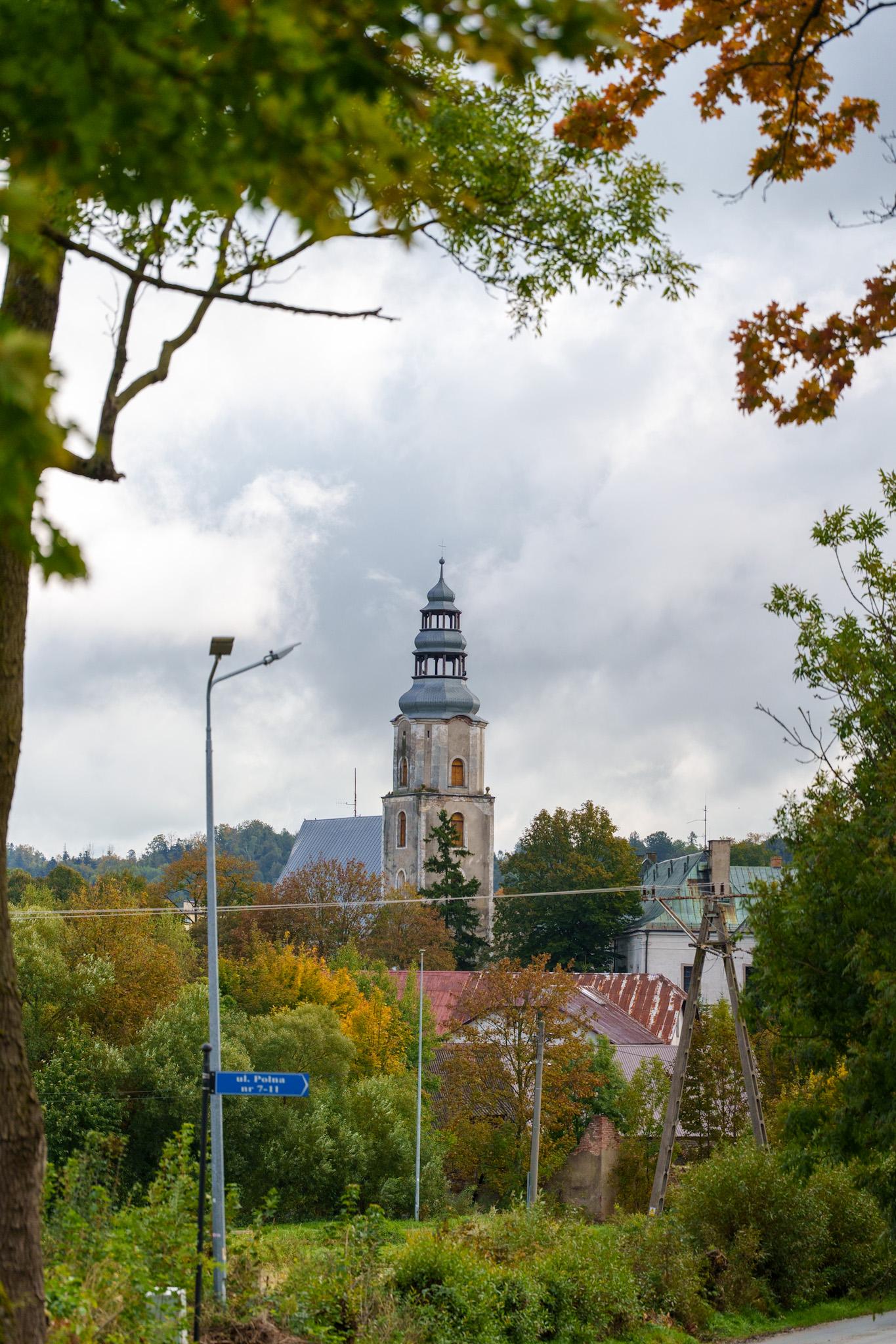 Malowniczy widok na wieżę kościelną wyłaniającą się spośród jesiennych drzew pod zachmurzonym niebem.