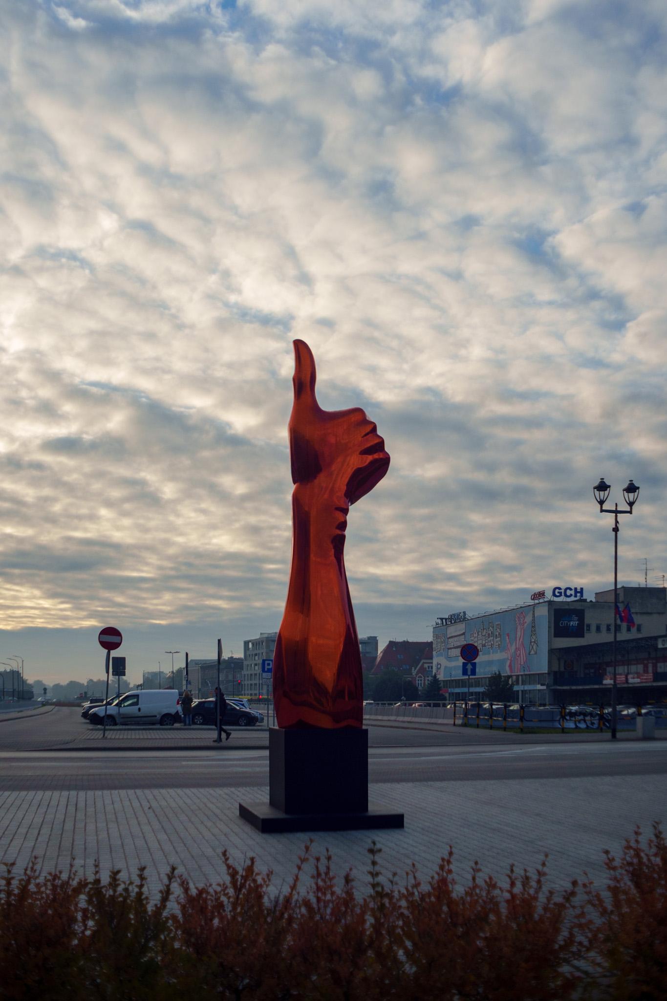 Nowoczesna rzeźba przedstawiająca gest kciuka w górę na tle malowniczego, zachmurzonego nieba o świcie. Industrialny krajobraz miasta z artystycznym akcentem.