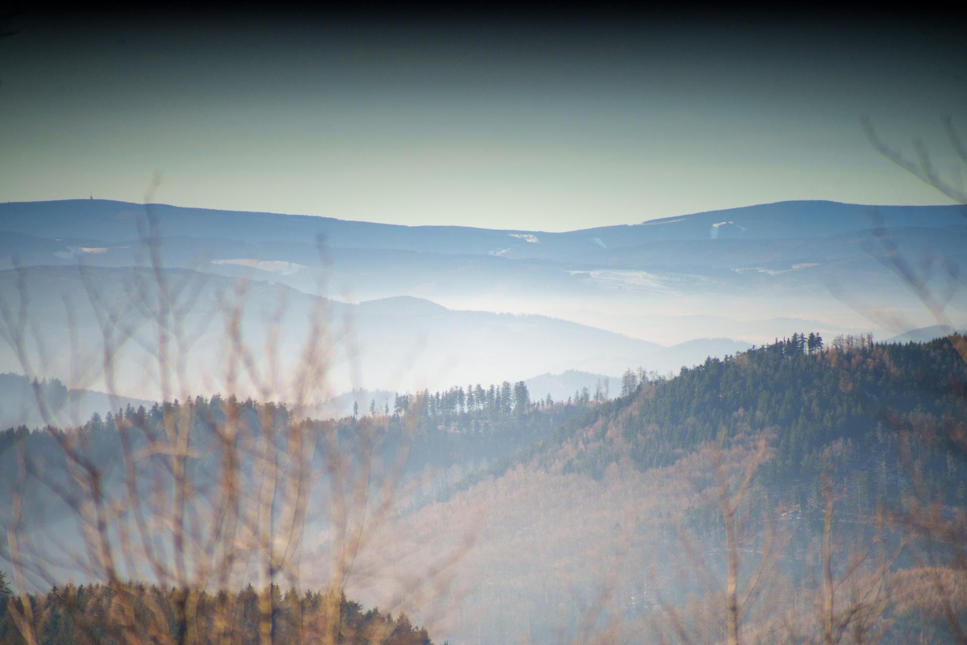 Malowniczy widok na zamglone pasma górskie o poranku, ukazujący warstwową strukturę krajobrazu z rozmytymi gałęziami na pierwszym planie.