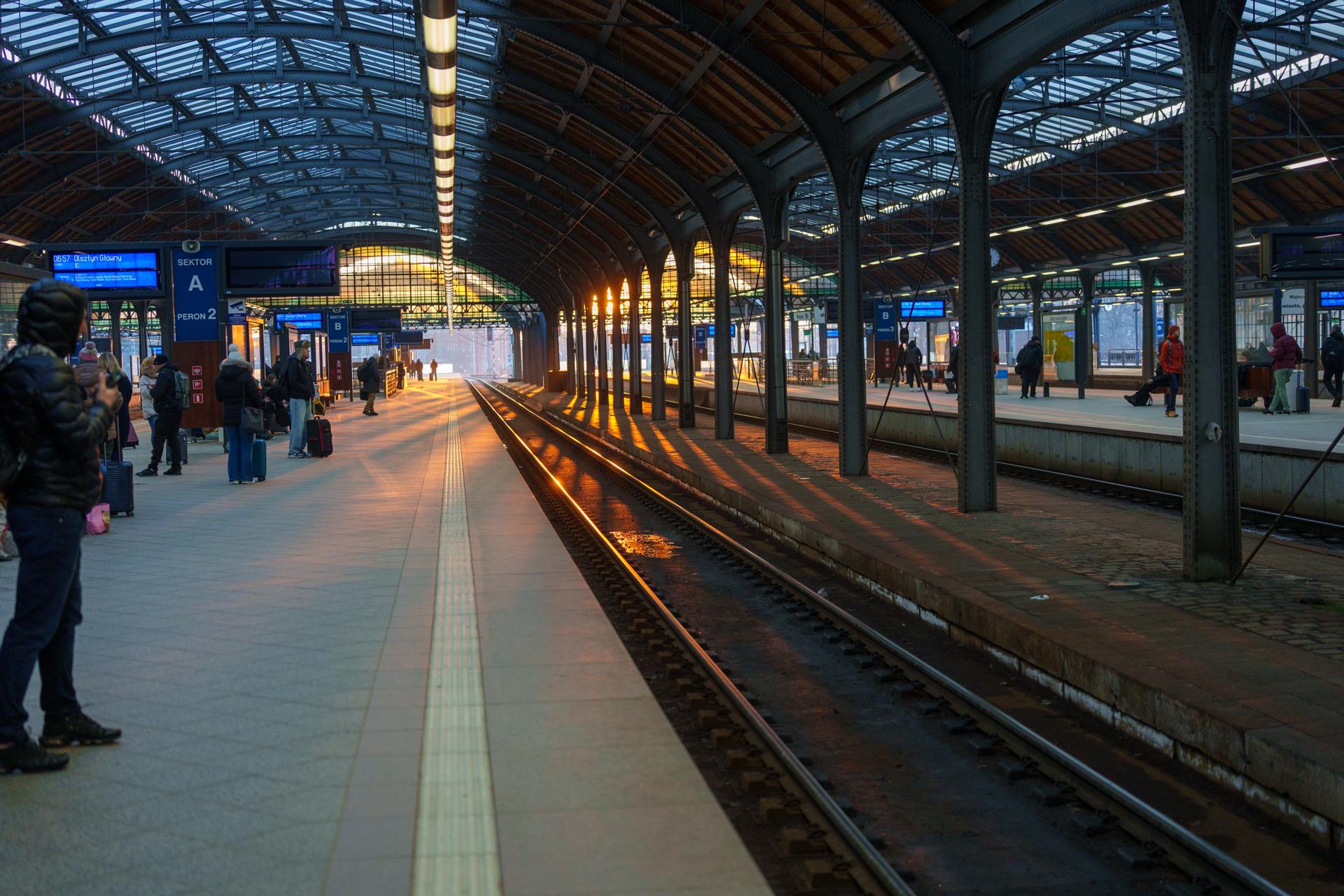 Malowniczy widok na peron dworca kolejowego o świcie, gdzie złote światło wschodzącego słońca odbija się od torów, tworząc nastrojową atmosferę porannej podróży.