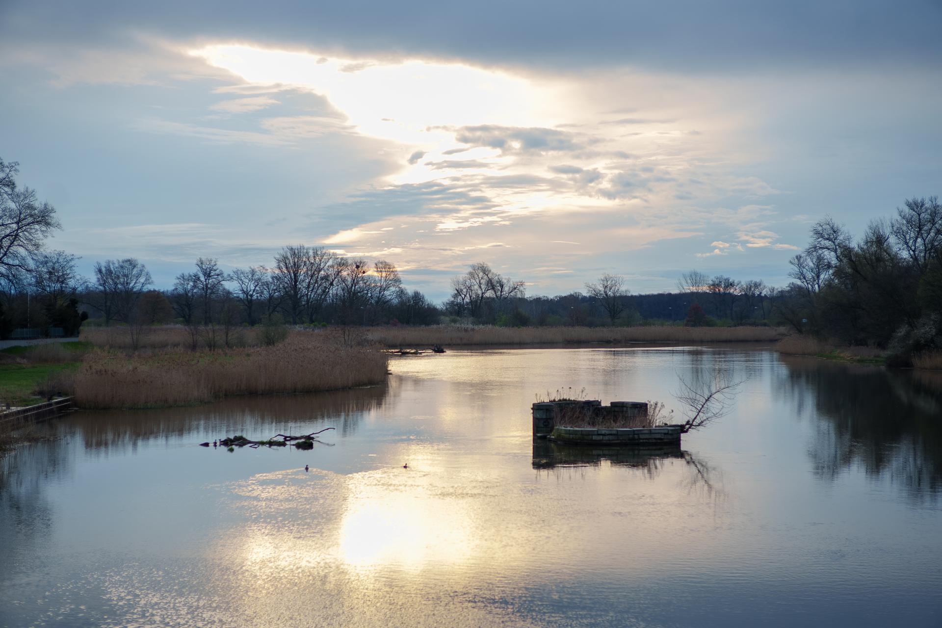 Malowniczy wschód słońca nad spokojną rzeką w wiosenny poranek. Słońce przebija się przez chmury, tworząc złociste refleksy na tafli wody.