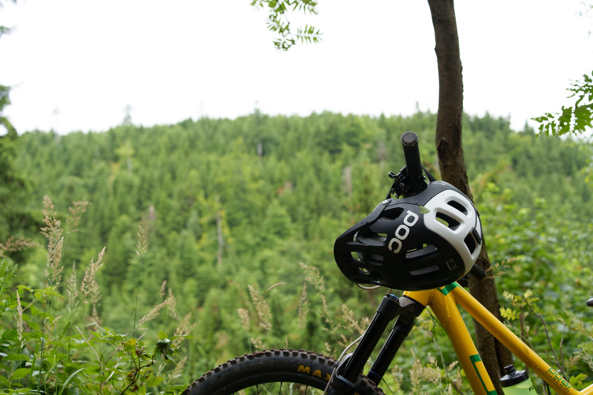 Żółty rower górski z kaskiem POC zawieszonym na kierownicy, uchwycony na tle gęstego, zielonego lasu. Idealny moment na odpoczynek podczas trailowej przygody.